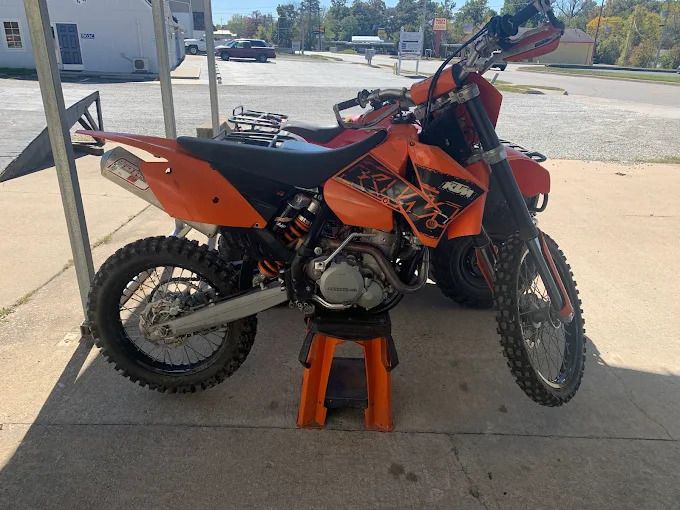 Orange dirt bike on a stand outside a building, parked on pavement in daylight.