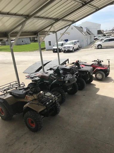 Three parked ATVs under a metal carport, with houses and a pickup truck in the background.