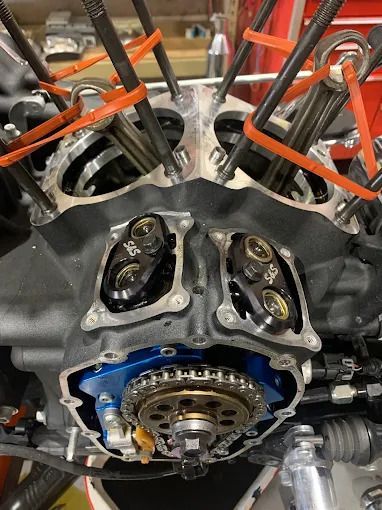 Close-up of a car engine cylinder head with exposed valves, orange hoses, and blue timing cover during repair