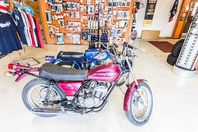 Pink and black vintage motorcycle displayed indoors in a shop with hanging clothes and wall art.