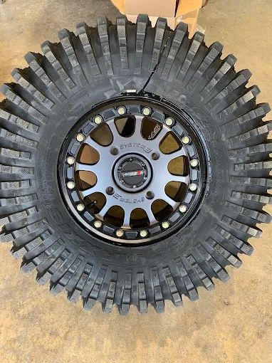 Off-road tire mounted on a black and silver wheel, viewed from above on a concrete floor