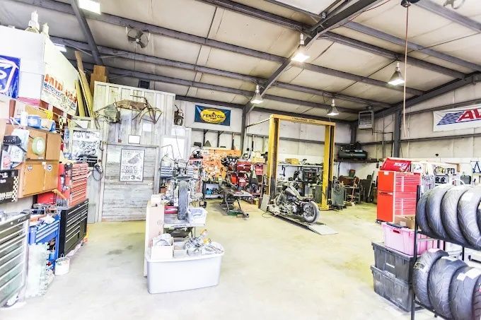 Auto repair shop interior with motorcycles, tires, tools, and shelves under a high metal ceiling