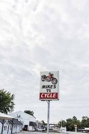Roadside sign for Mike's 71 Cycle with a red motorcycle logo against a cloudy sky