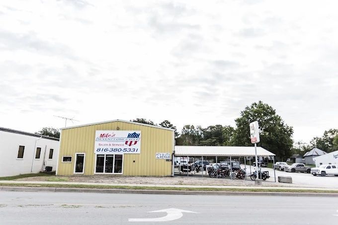 Small auto dealership lot with a yellow office building, carport, and parked cars under a cloudy sky