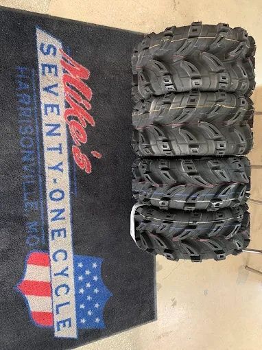 Stack of black off-road tires beside a “Seventy-One Cycle” floor mat with red and blue graphic design.