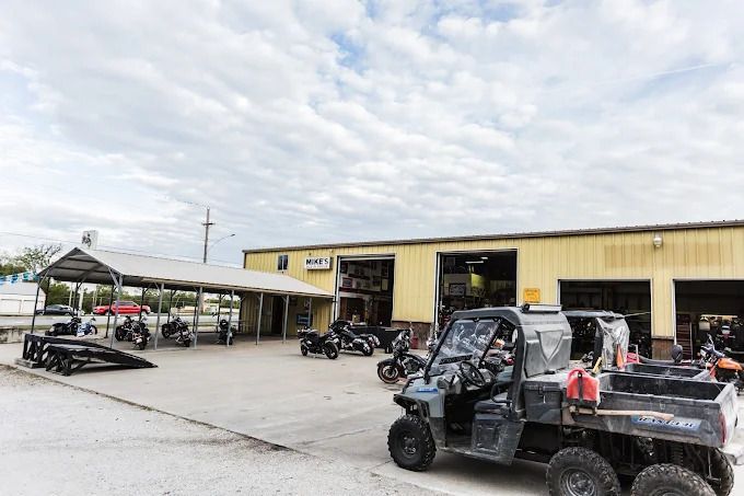 Open-air repair shop with ATVs and a black utility vehicle parked outside a yellow building.