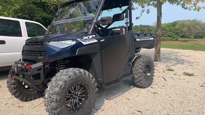 Black off-road UTV parked on a gravel lot beside a white van, with trees in the background