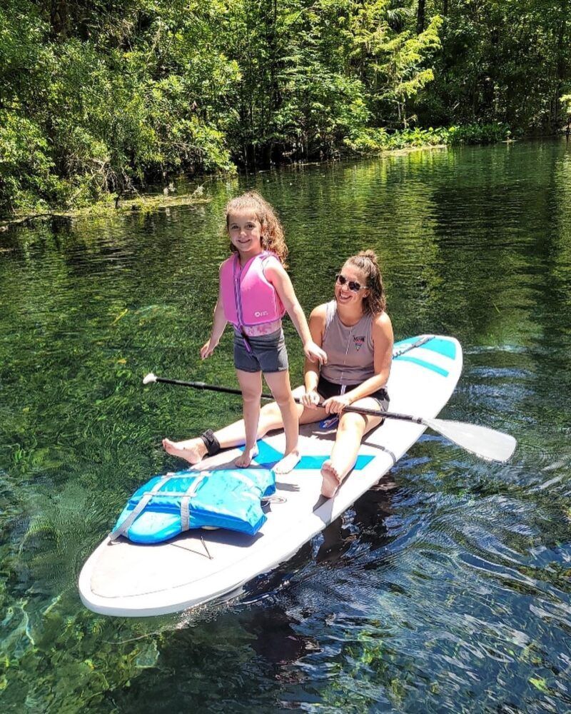 Woman and child paddleboarding on clear water, girl wearing a pink life vest.
