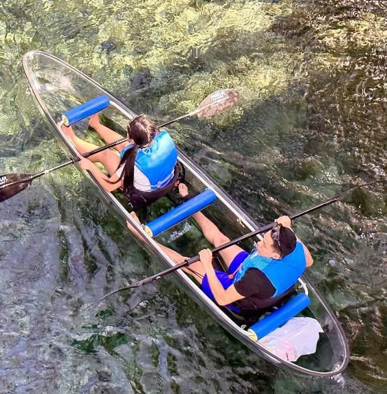 Two people kayaking in a clear canoe on clear water. They are wearing life vests and using paddles.