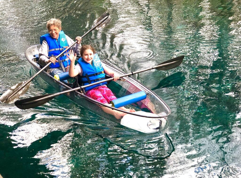 Two people in a clear kayak paddle on water, one waves.