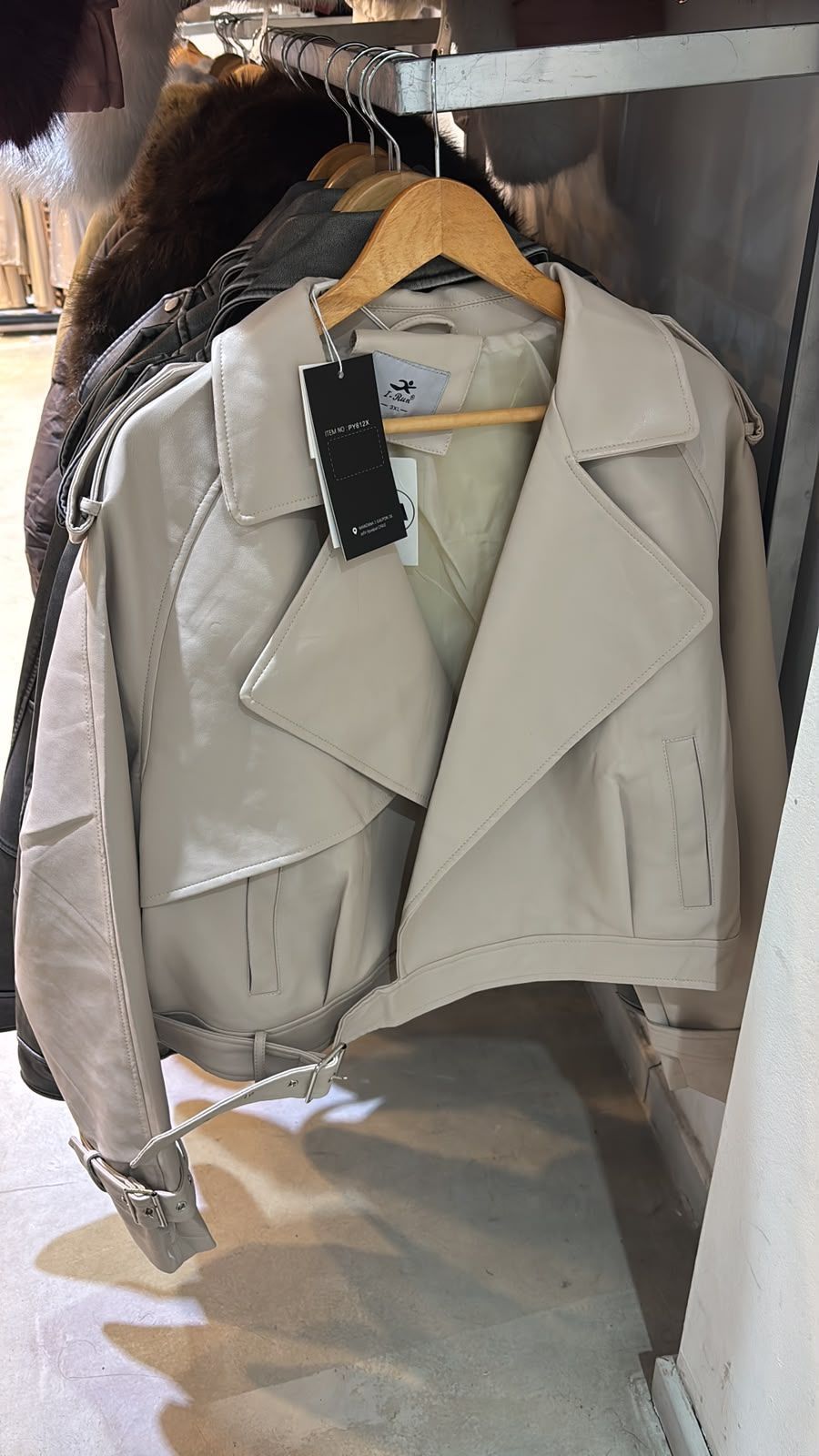 Chaqueta color beige claro con una percha y etiqueta negra colgada en un armario o perchero.
