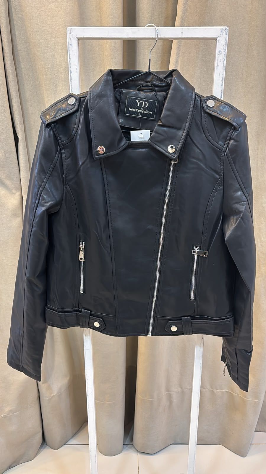 Chaqueta de cuero negra de motorista colgada en un perchero blanco frente a unas cortinas beige.