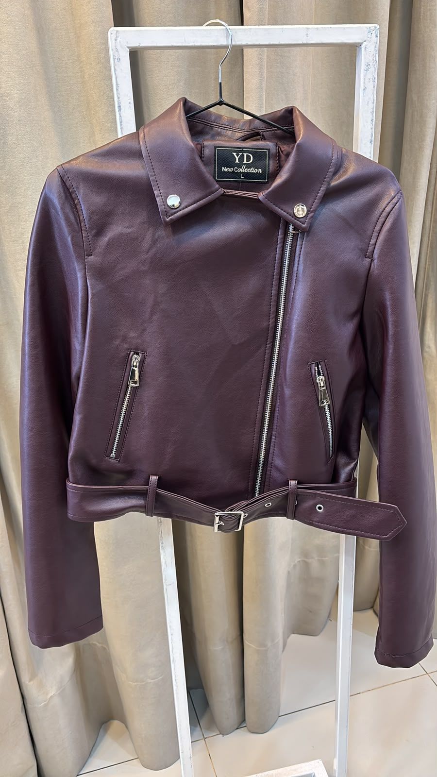 Chaqueta de cuero morada con cremallera y cinturón asimétricos, colgada sobre una cortina.