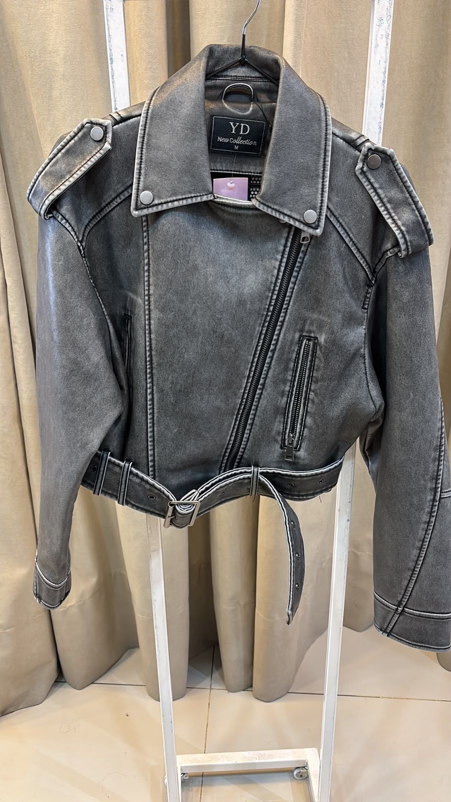 Chaqueta corta de mezclilla gris colgada en una percha frente a cortinas beige.