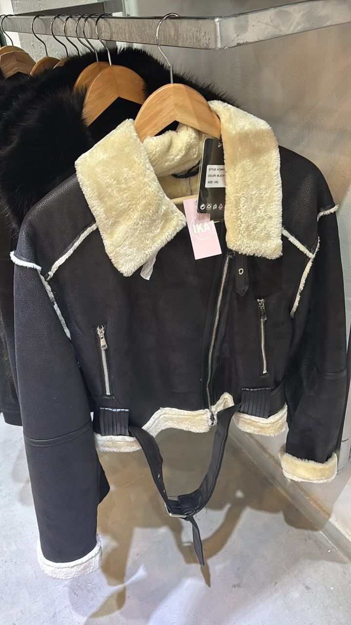 Chaqueta negra de borrego con cuello y puños color crema, colgada en un perchero junto con otros abrigos.