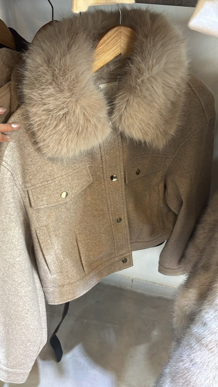 Abrigo color topo con un gran cuello de piel colgado en un perchero, visto desde el frente.
