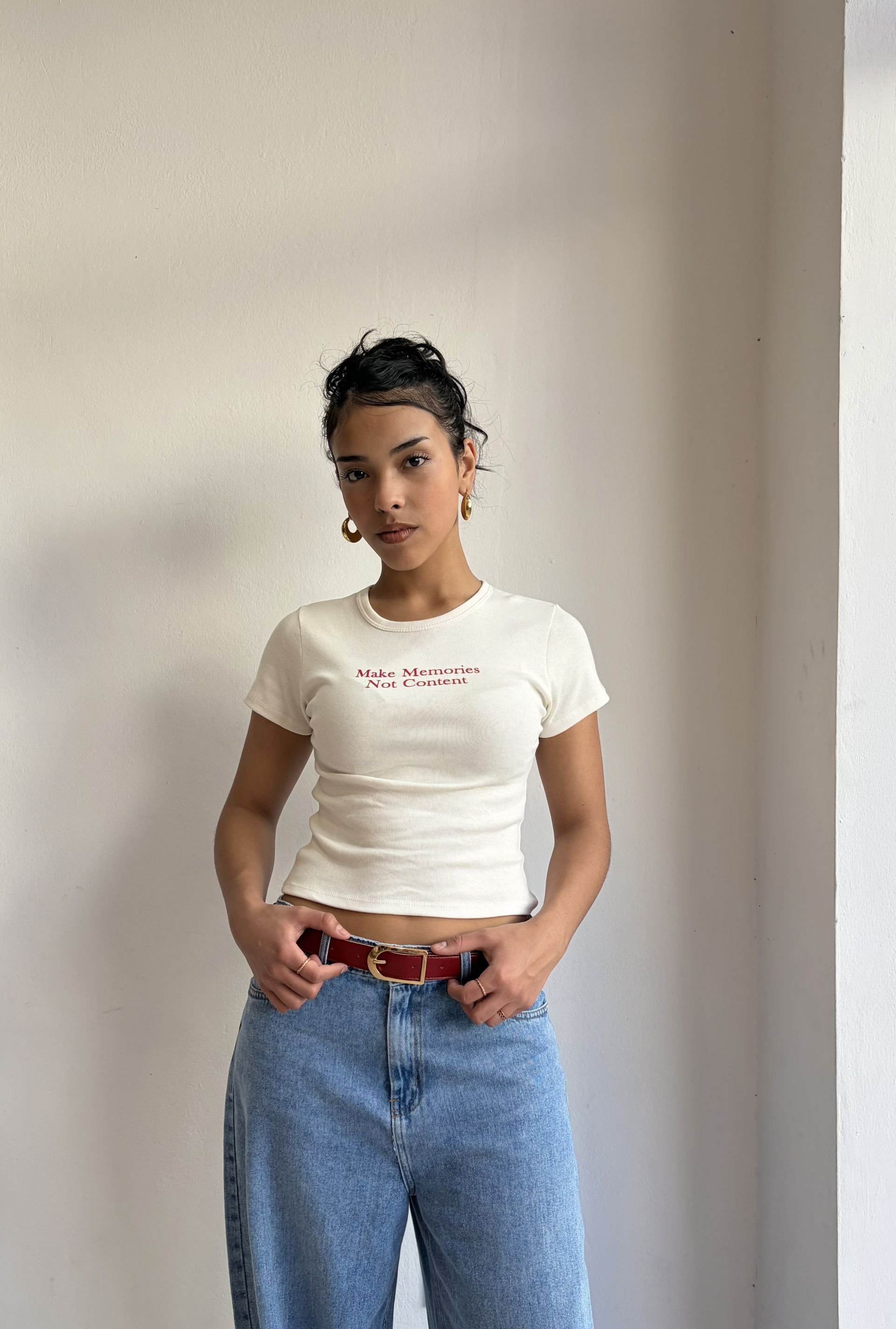 Persona con remera blanca y letras rojas con un jeans azul contiene un cinto bordo. posando al frente de la pared.