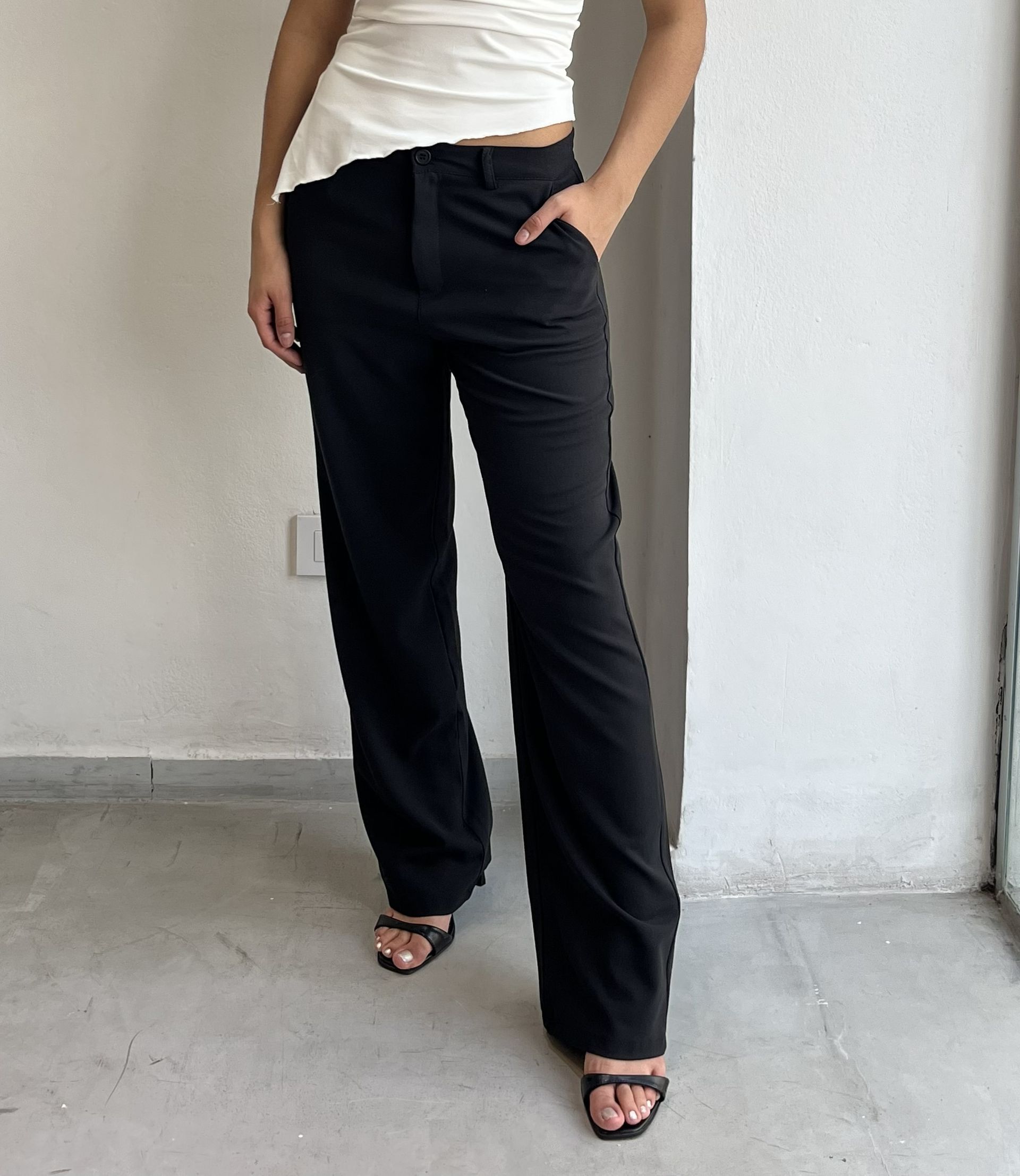 Persona posando con un pantalón negro y sandalias negras adelante de una pared blanca.