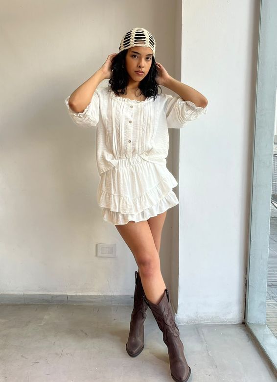 Persona posando con un suéter de rayas, falda negra y botas frente a unas cortinas grises.