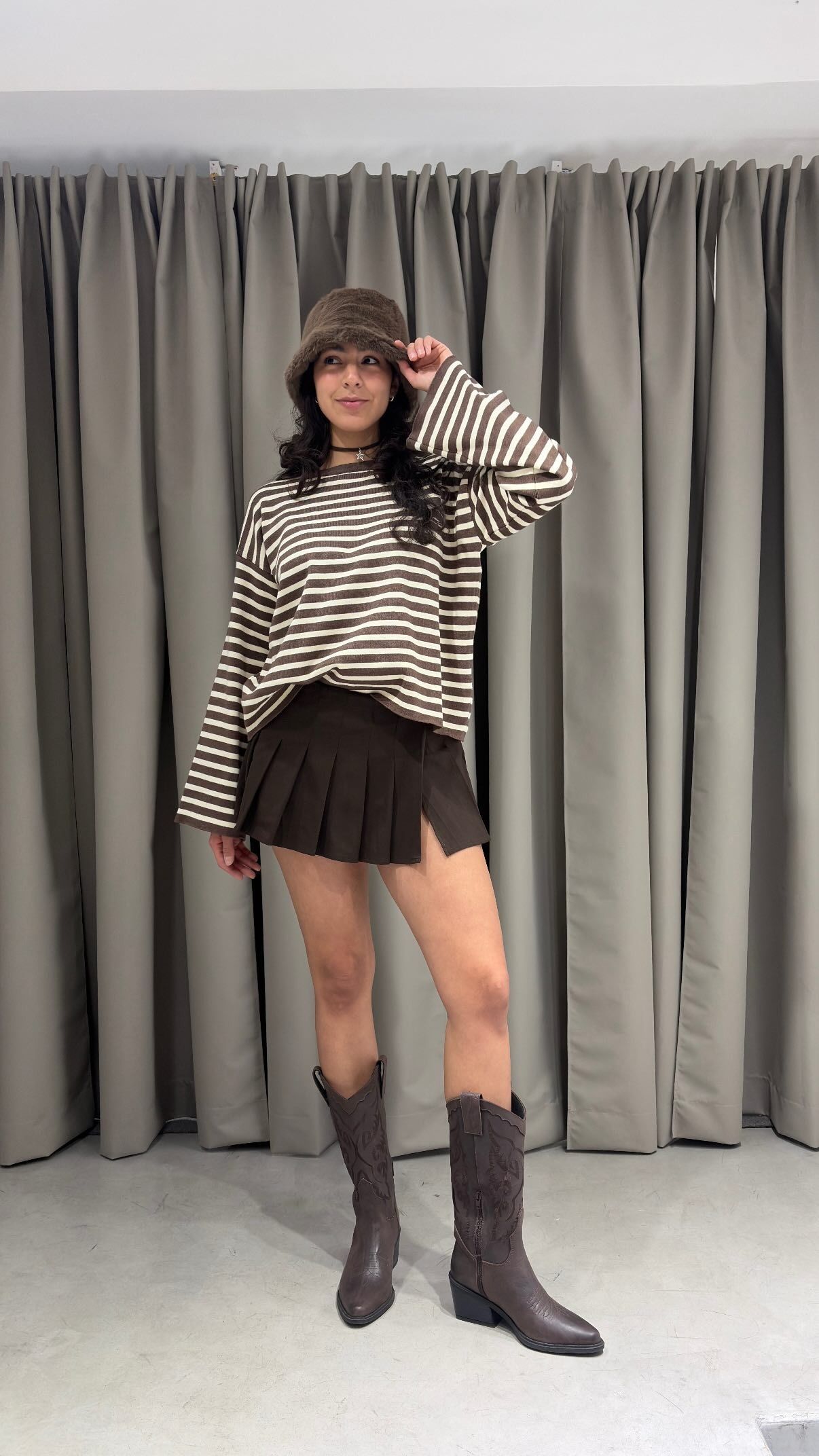 Persona con suéter de rayas, falda negra y botas altas posando frente a unas cortinas grises.