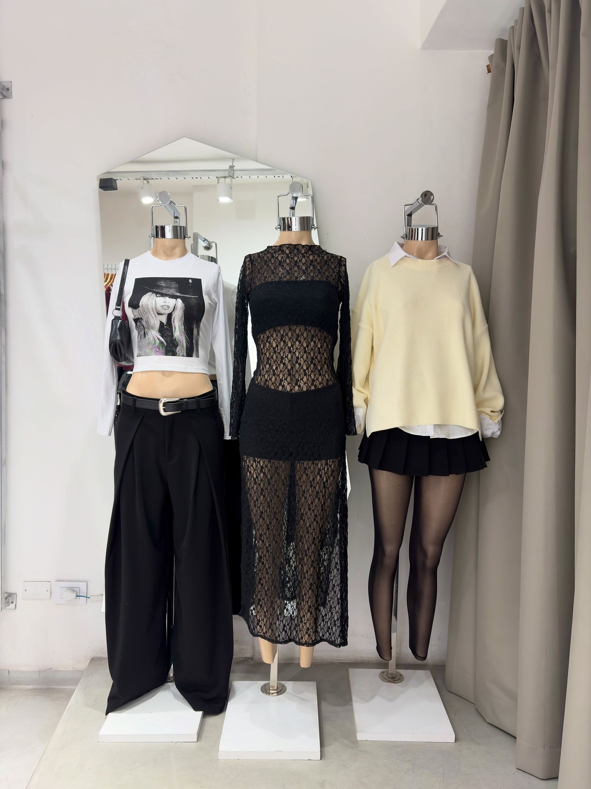 Tres maniquíes con atuendos negros y crema se encuentran en una habitación luminosa cerca de un espejo y una cortina.
