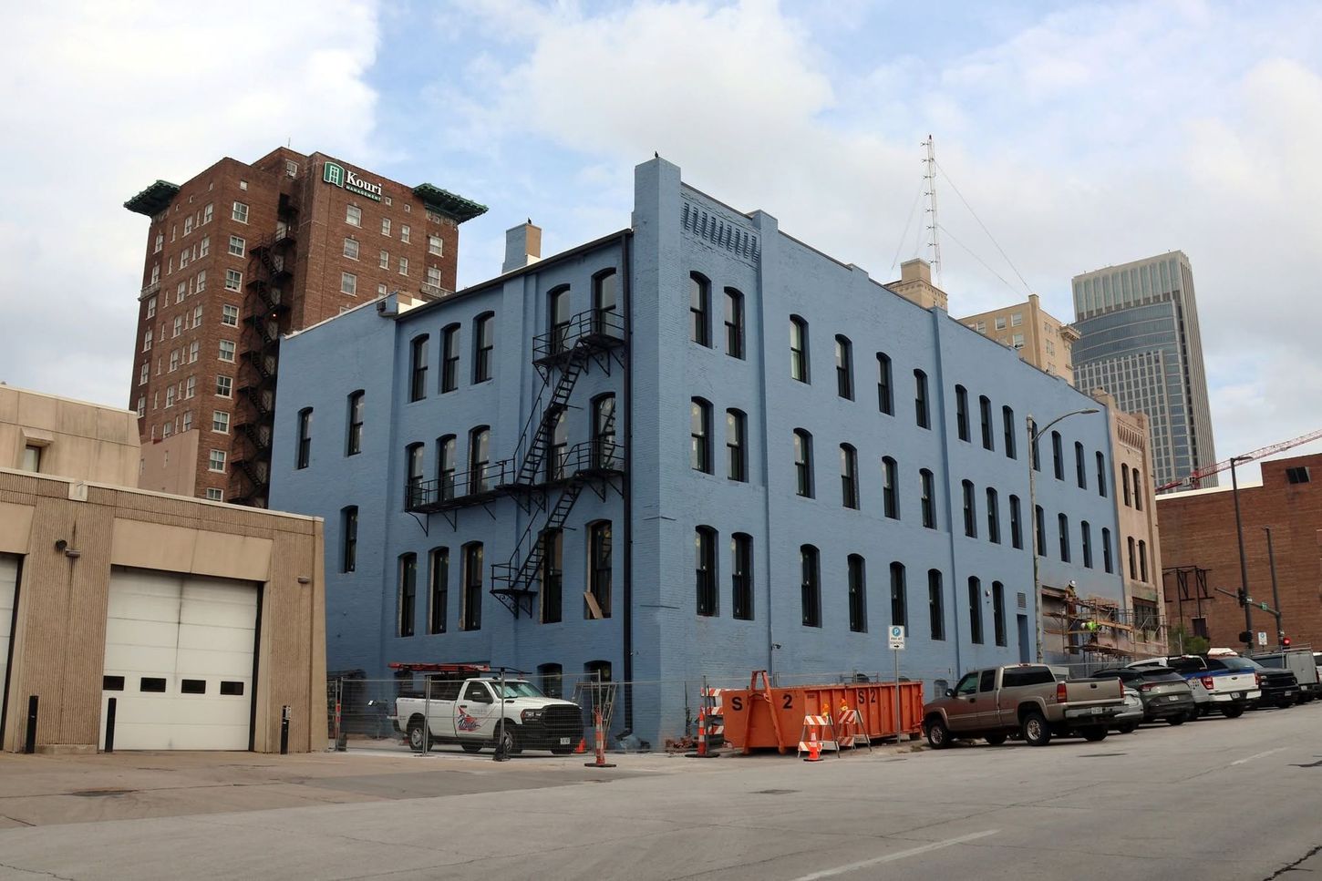 Blue industrial building on a city street with parked cars and an orange dumpster under a cloudy sky