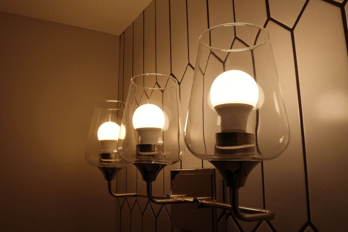 Three glowing white bulbs in clear glass wall sconces against a warm beige wall.