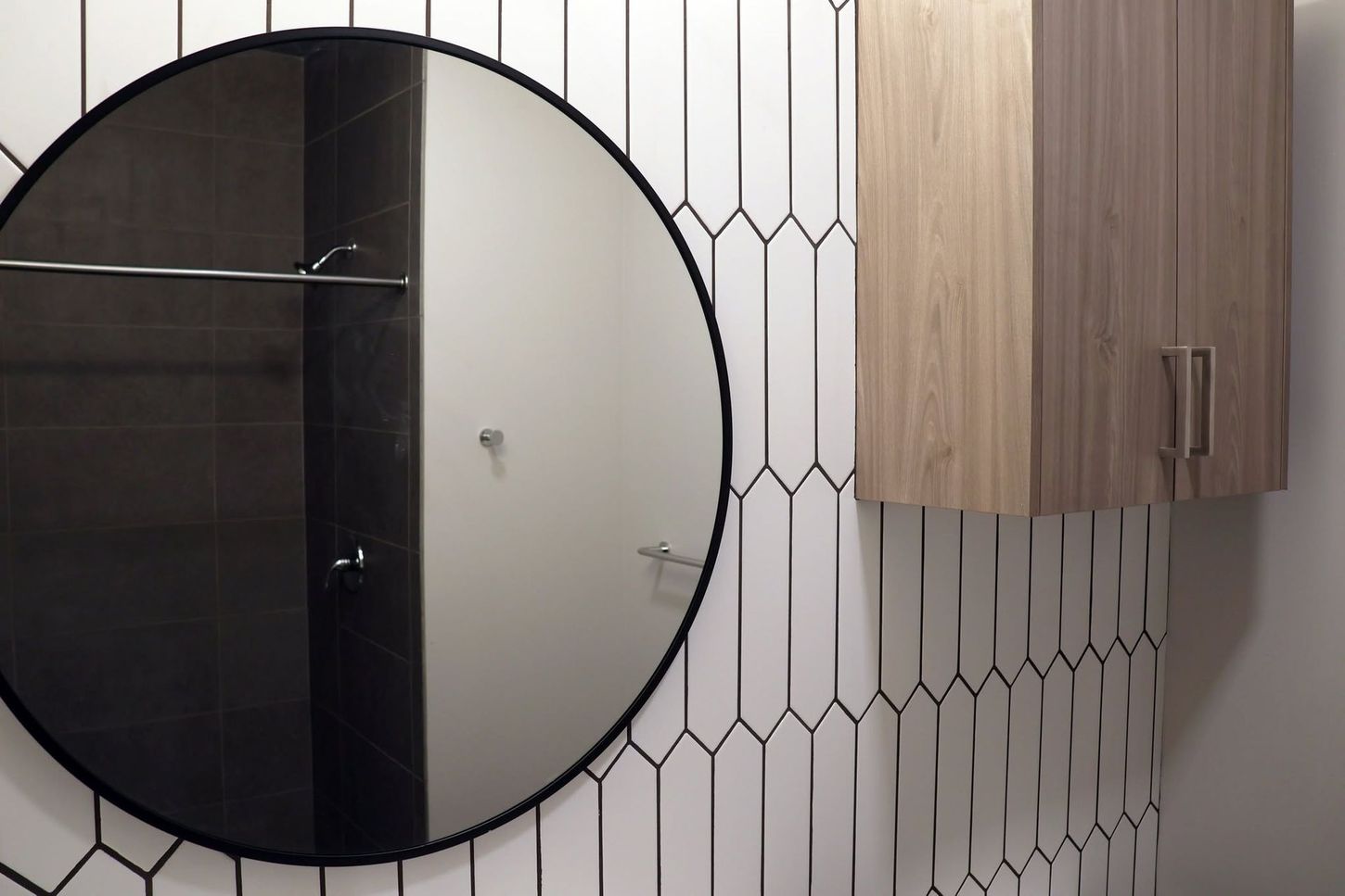 Round black-framed mirror on a tiled bathroom wall beside a light wood cabinet