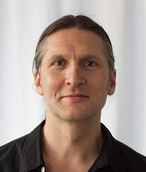 Man with long gray hair, wearing a black shirt, smiling.