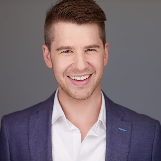 Man with short brown hair smiles, wearing a blue blazer over a white shirt, against a gray background.