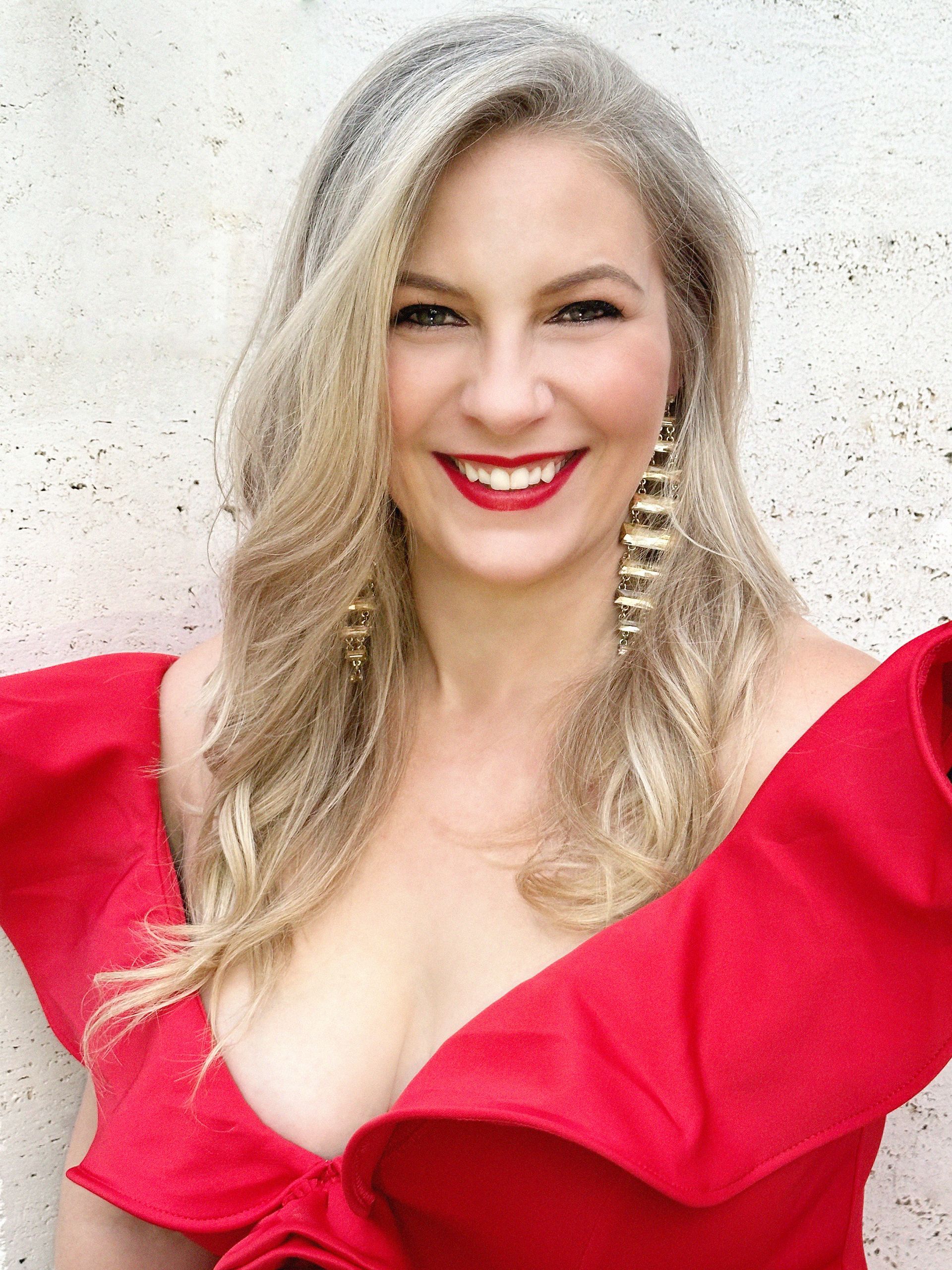 Woman with blonde hair, red dress, and large gold earrings smiling in front of a white wall.