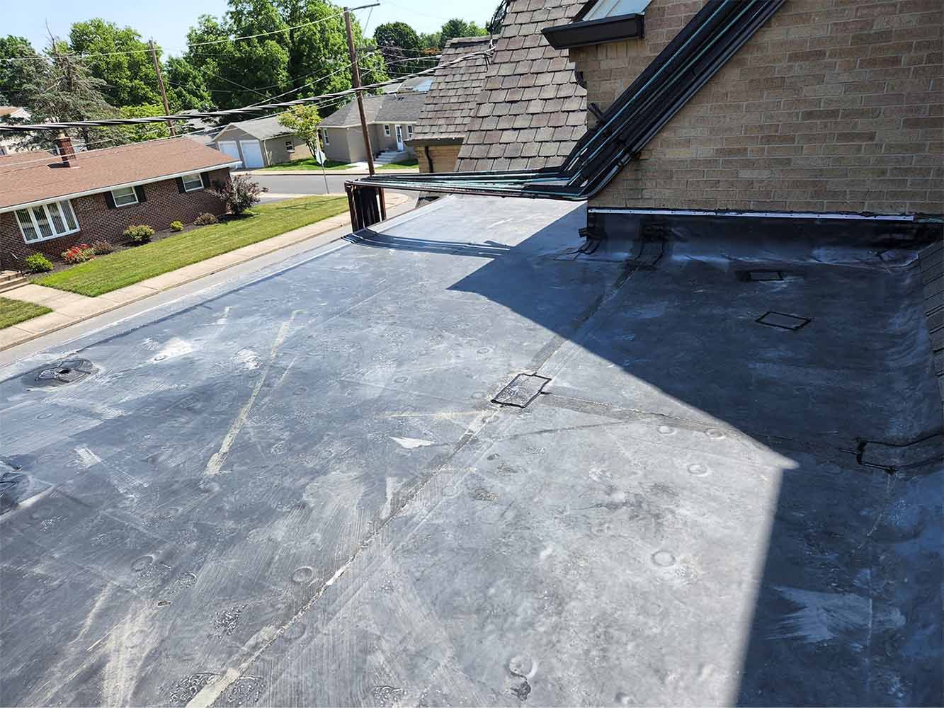 Flat rooftop with black roofing membrane beside a sloped shingle roof, overlooking a suburban neighborhood.