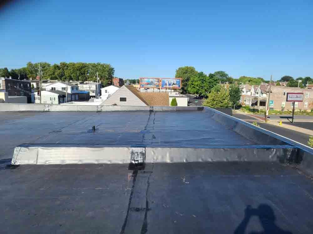 Flat rooftop with black waterproof membrane overlooking trees, houses, and a clear blue sky.