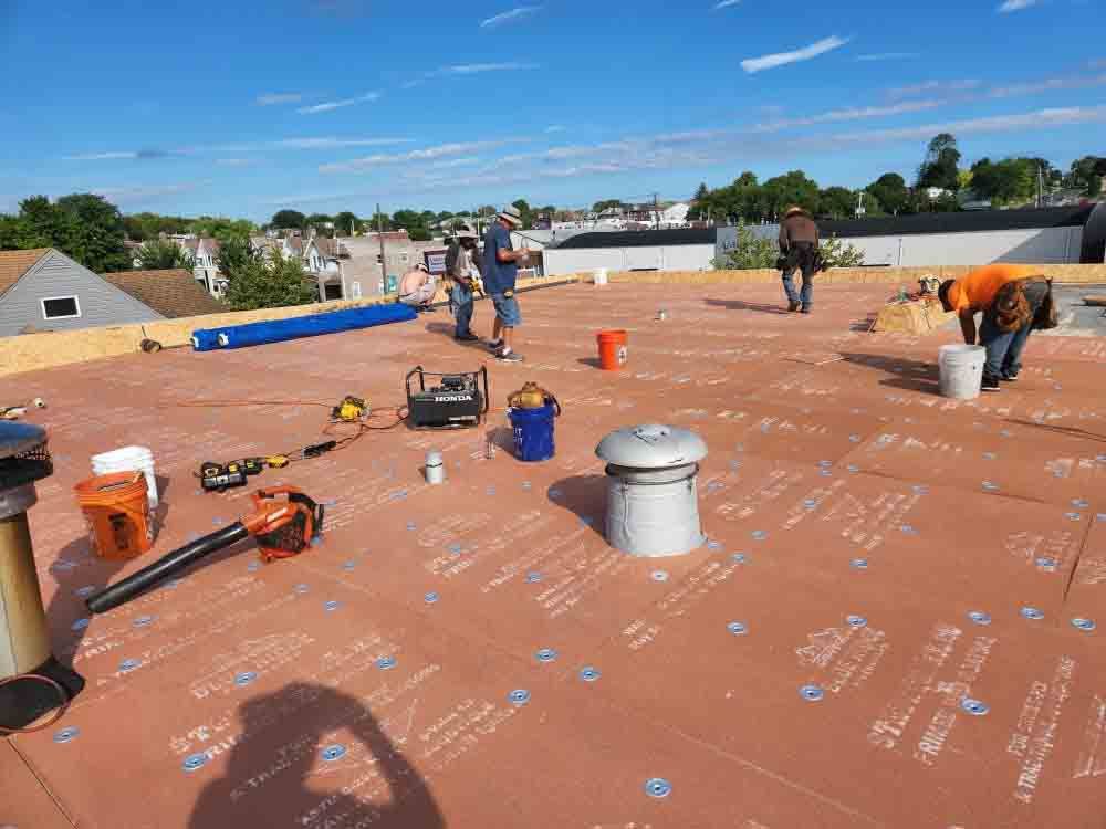 Workers installing roofing on a flat rooftop with tools, buckets, and orange safety cones under a blue sky