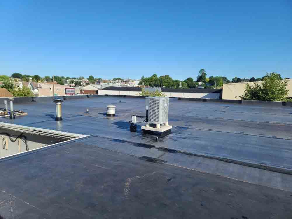 Flat rooftop with black waterproofing, vents, and a clear blue sky over nearby buildings