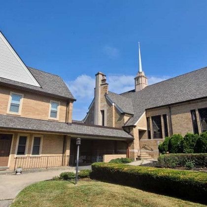 Courtyard of a brick church with a gray roof and tall white steeple under a clear blue sky