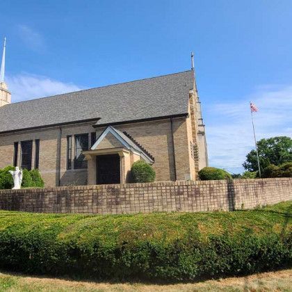 Stone church with a steep roof and small entrance porch, behind a low stone wall and green shrubs under a blue sky