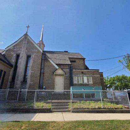 Stone church behind a chain-link fence under a blue sky, with a sidewalk in front.