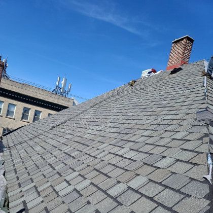Steep gray shingled roof against a blue sky, with a brick chimney and nearby building windows.