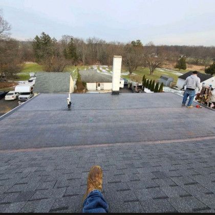 View from a flat shingled roof with workers near the edge and a rural neighborhood beyond