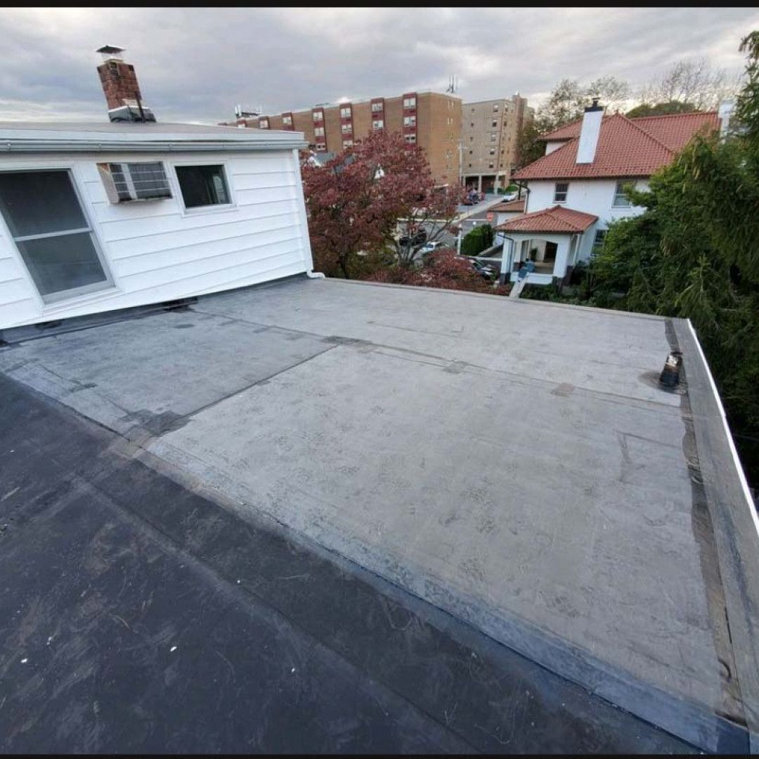 Flat rooftop with gray roofing, adjacent white house, and trees and apartment buildings in the background.