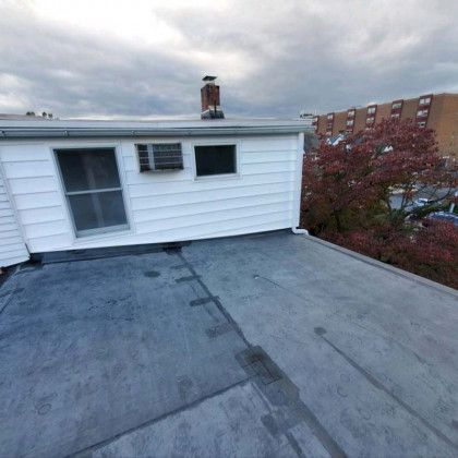 Flat rooftop with gray membrane, white siding, windows, and a chimney under a cloudy sky.