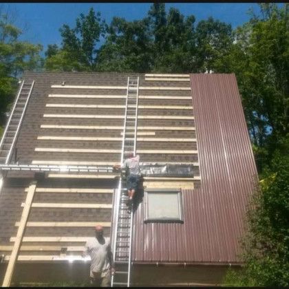 Two workers on ladders installing wood slats on a cabin roof exterior under trees
