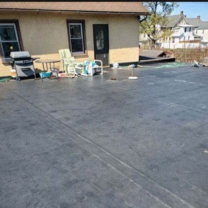 Flat rooftop patio beside a house, with chairs, grill, and stored items under a clear sky.