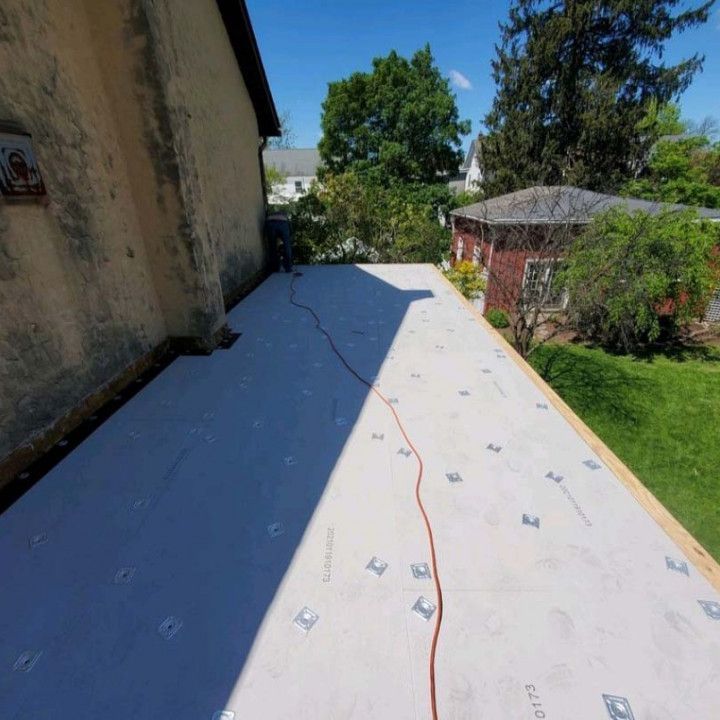 Flat white roof under construction beside a house, with red cable and trees in the background