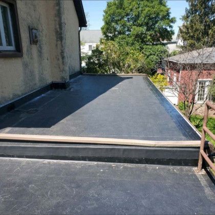 Flat black rooftop or patio beside a stucco house, with trees and neighboring buildings in the background.