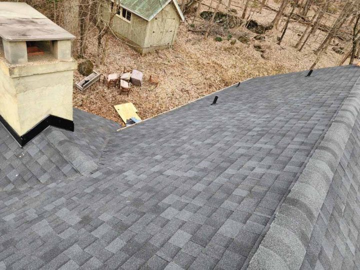 Roof with gray shingles and a chimney, overlooking a wooded yard with small sheds.