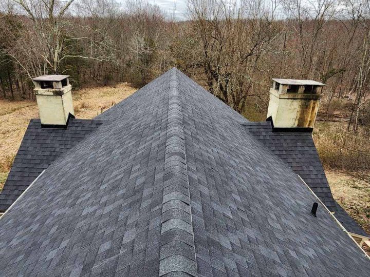 Roof with dark gray shingles and two chimneys, viewed from above in a wooded area.