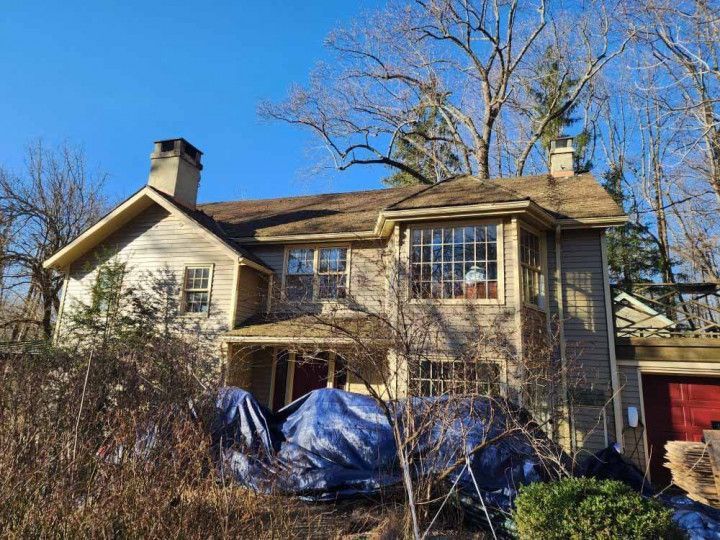 Two-story tan house with a blue tarp over the front porch, leafless trees, and a red garage on the right.