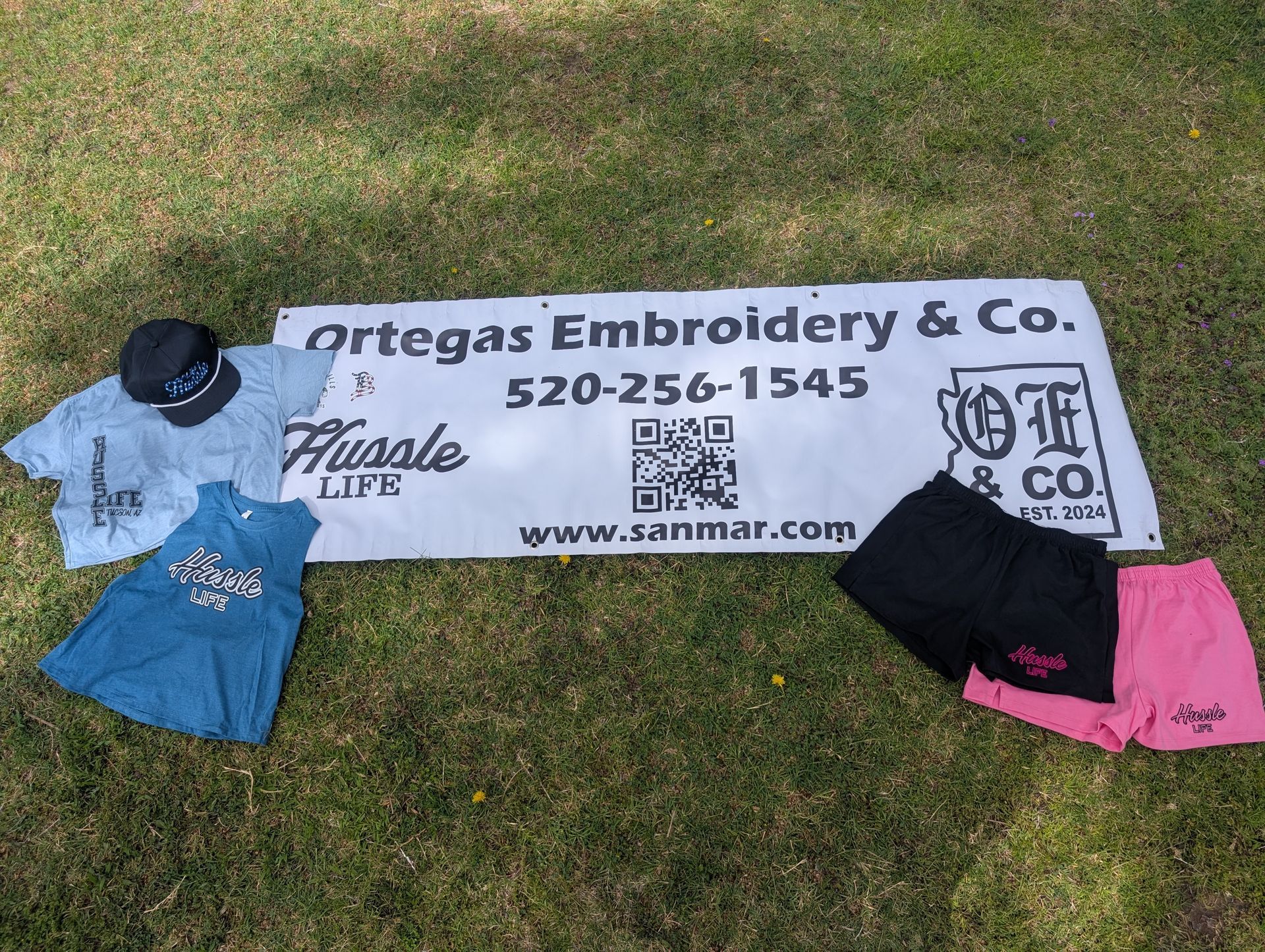 A sign for ortegas embroidery & co. is sitting on top of a lush green field.